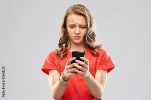 technology, cyberbullying and emotions concept - sad teenage girl in red t-shirt using smartphone over grey background
