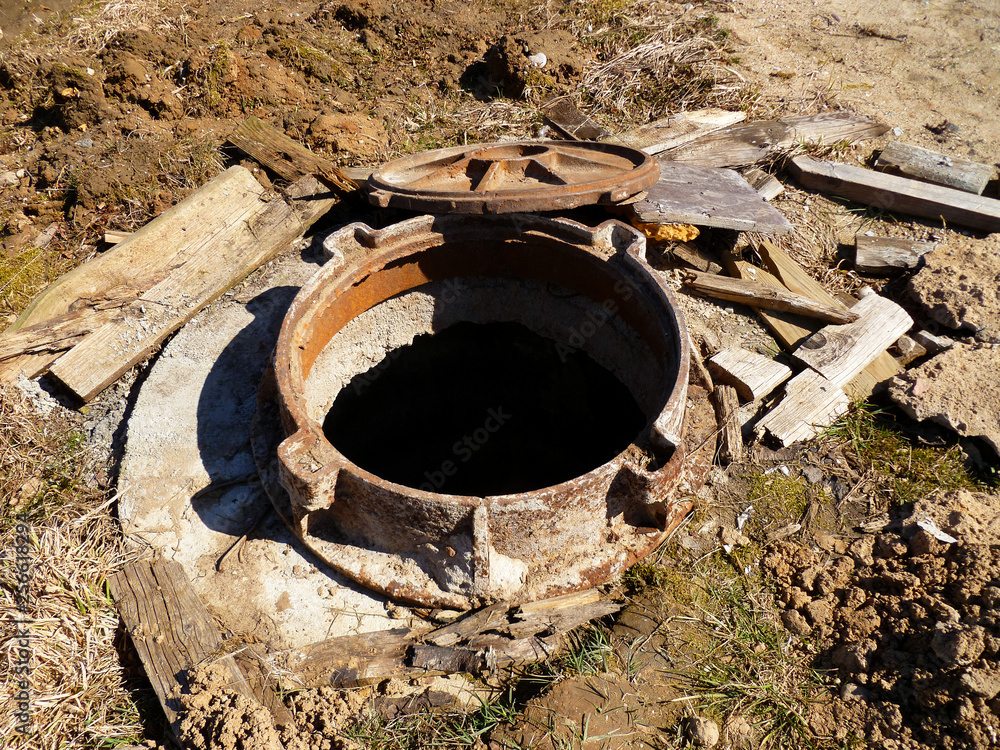 Sewerage hatchway, manhole. Rusty pit cover ring on construction site. Old cast rust iron sewerage hatchway with the growing grass and dirt around, closeup.