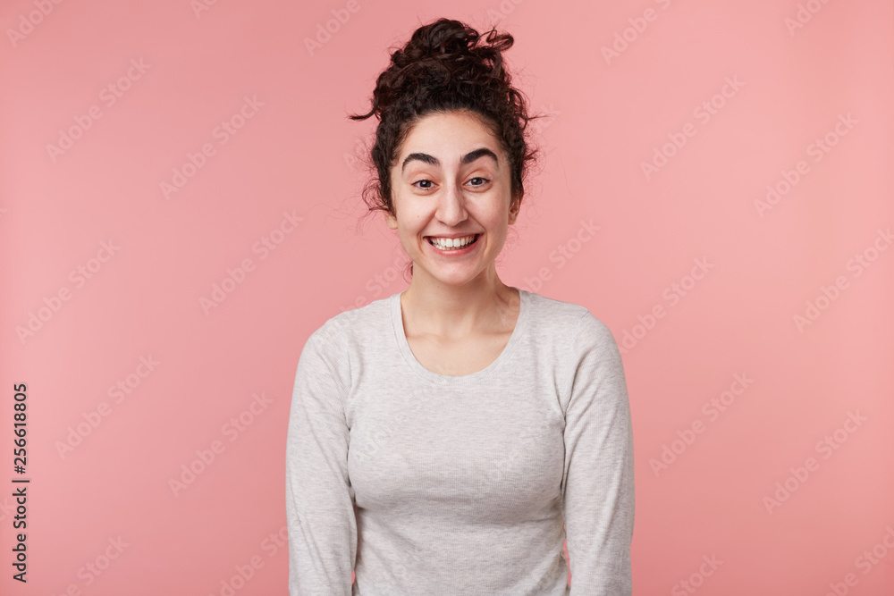 Close up of young women with dark curly hair, with closed eyes, wears casual white longsleeve, smiles broadly, isolated over pink background. Positive emotion concept.