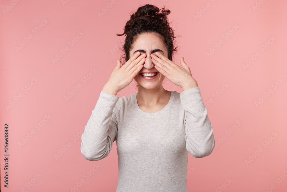 Close-up shot of beautiful young female with dark curly hair, wears casual white longsleeve, covering her eyes with hands, isolated on over pink background. Close-up shot of beautiful young female with dark curly hair, wears casual white longsleeve, covering her eyes with hands, isolated on over pink background.