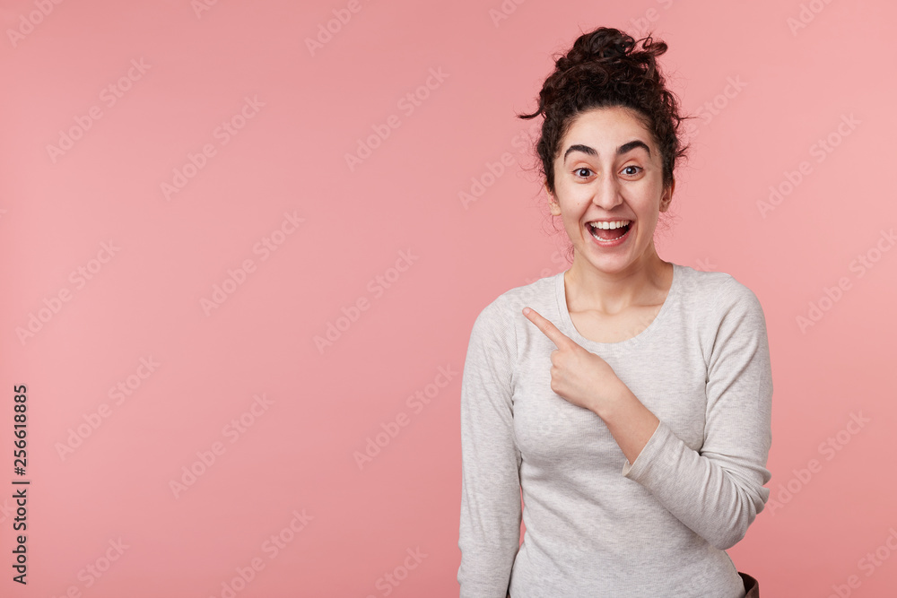 Fototapeta premium Photo of happy young woman standing isolated over pink background with copyspace for your advertising, points with one finger up, wears casual white longsleeve.