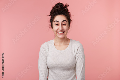 Close up of young women with dark curly hair, with closed eyes, wears casual white longsleeve, smiles broadly, isolated over pink background. Positive emotion concept. © timtimphoto