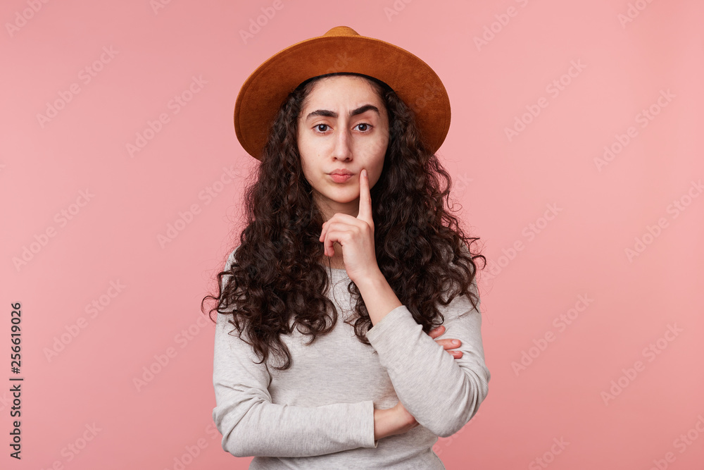 Close up of young lady with a hat on head, one finger touches cheek, standing isolated over pink background, looked up and thinking. Close up of young lady with a hat on head, one finger touches cheek, standing isolated over pink background, looked up and thinking.