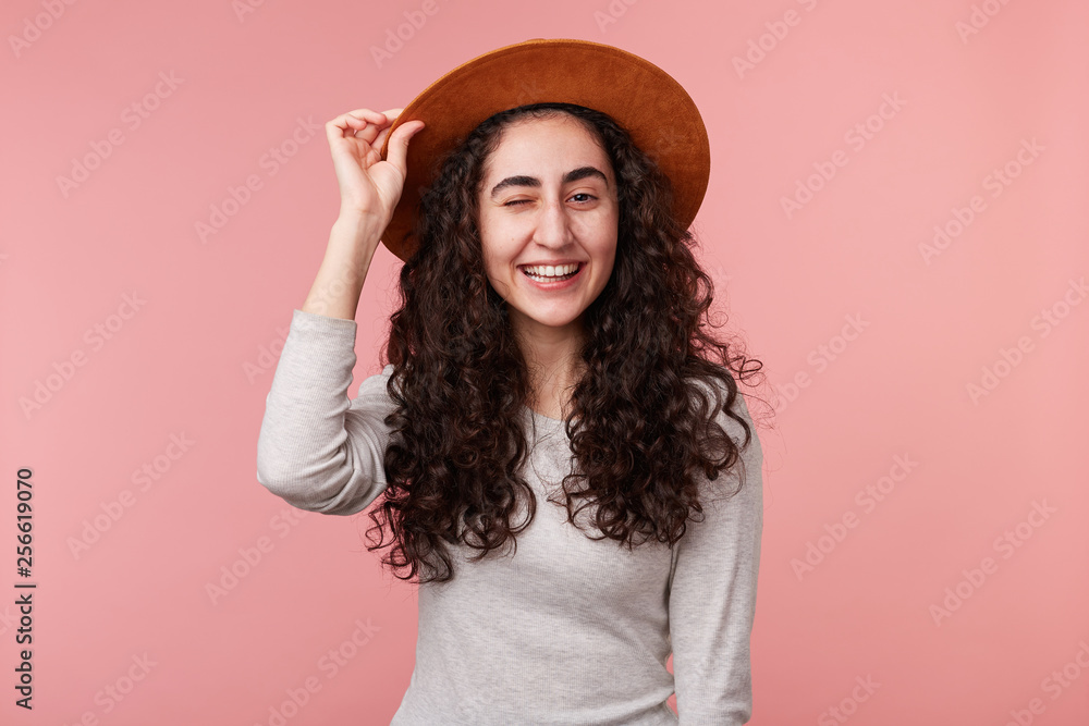 Photo of happy young woman with curly hair, holds hat on head and winking, wears casual white longsleeve, isolated over pink background. Positive emotion concept. Photo of happy young woman with curly hair, holds hat on head and winking, wears casual white longsleeve, isolated over pink background. Positive emotion concept.