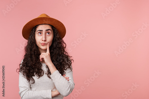 Image of thinking young lady with a hat on head, one finger touches cheek, standing isolated over pink background, looked up, with copy space for your text or advertising content.