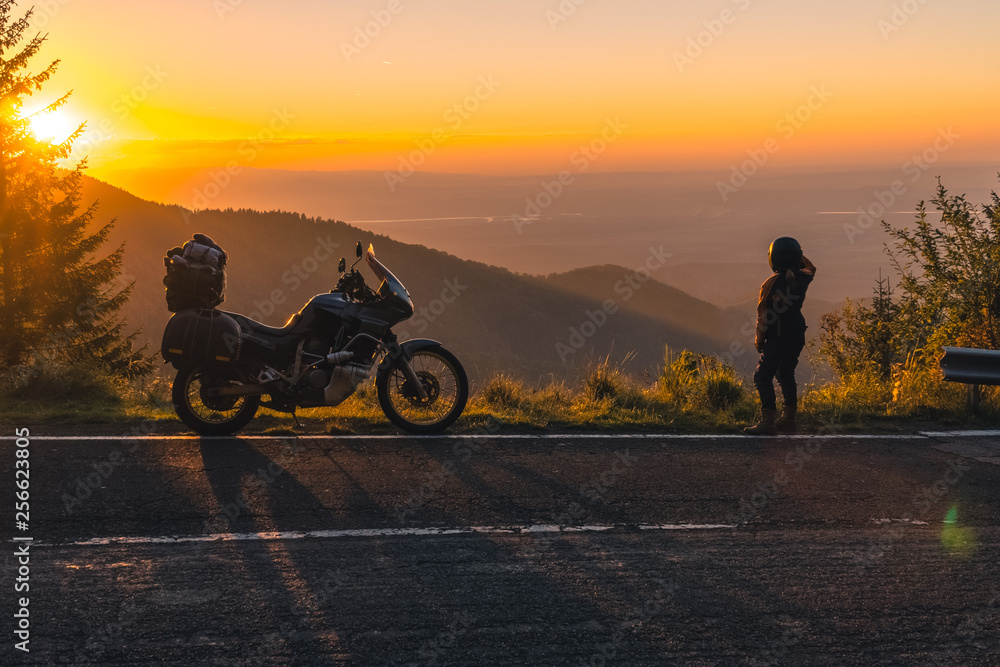 girl in full motorcycle equipment, stands on the side of the road over a cliff and looks into the distance at beautiful sunset in the mountains. Adventure motorcycle, Transfagarasan, Romania
