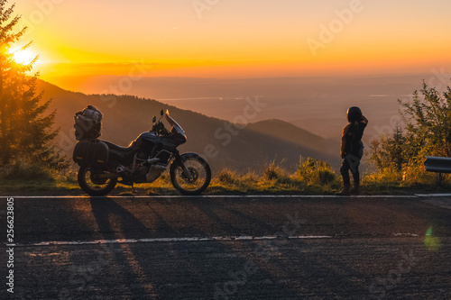 girl in full motorcycle equipment, stands on the side of the road over a cliff and looks into the distance at beautiful sunset in the mountains. Adventure motorcycle, Transfagarasan, Romania photo