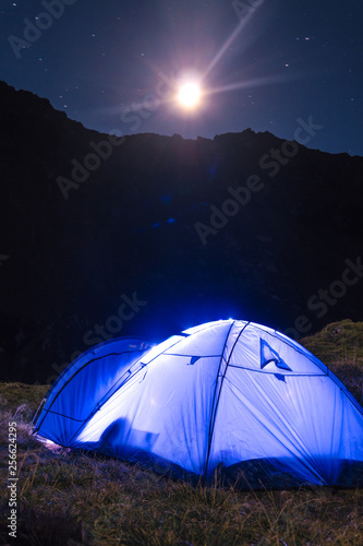 Night mountain landscape with illuminated blue tent. Mountain peaks and the moon. outdoor at Lacul Balea Lake, Transfagarasan, Romania. Concept Travel, copy space, vertical photo