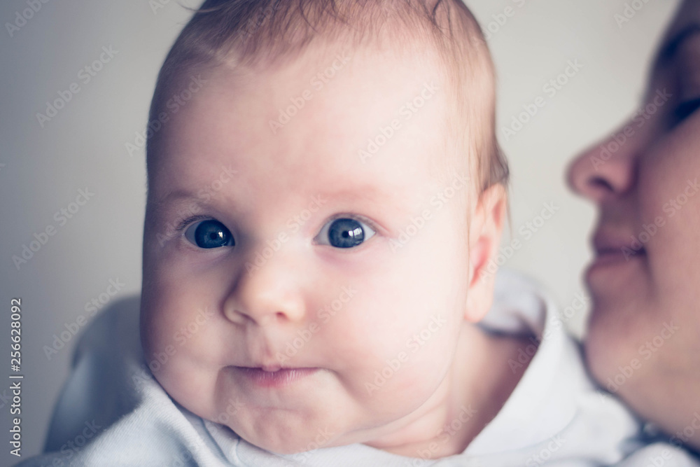 Mum kisses a baby baby in a cheek. The girl has a funny look. Caucasian child, blue eyes and red hair. Mom is not in focus.