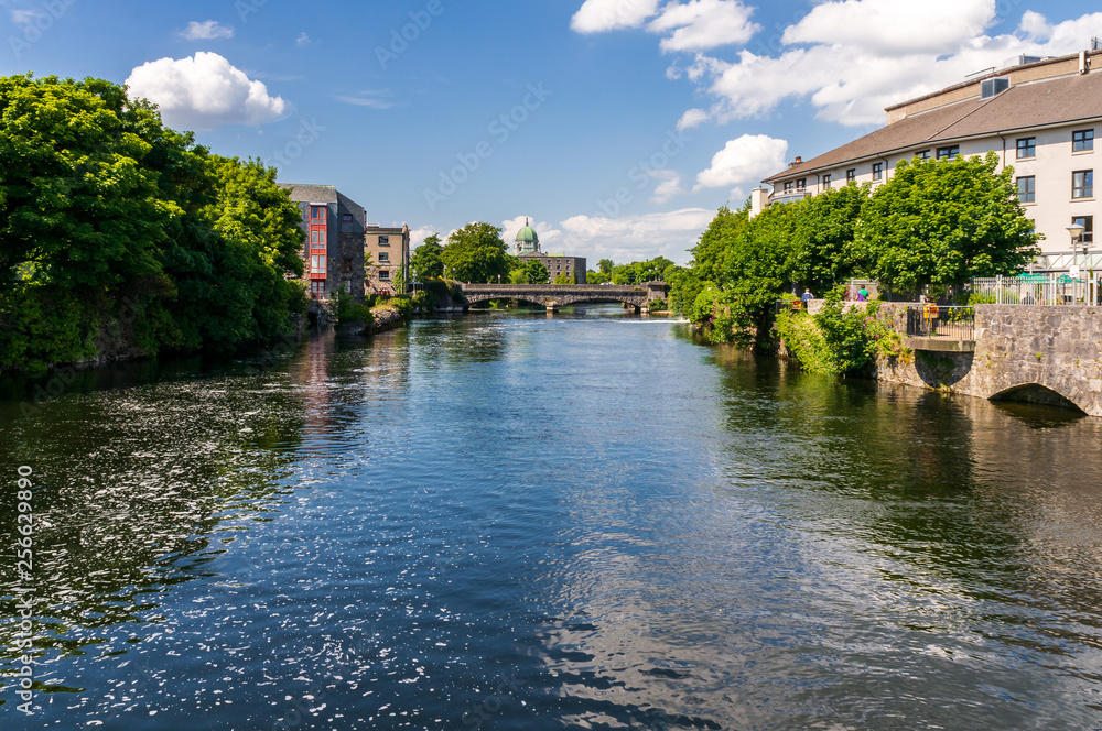 River Corrib landscape in Galway, Ireland, as seen from the Wolfe Tone Bridge under a beautiful blue sky on a sunny summer day, bordered by houses and  green trees.