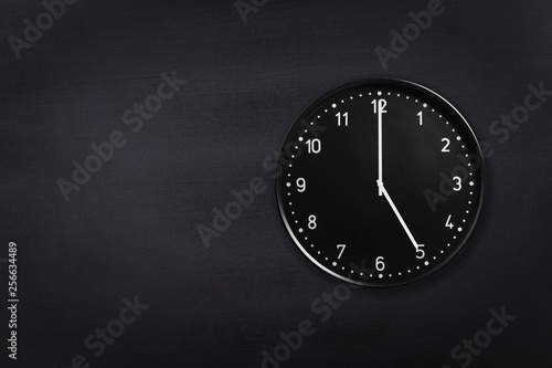 Black wall clock showing five o'clock on black chalkboard background. Office clock showing 5am or 5pm on black texture