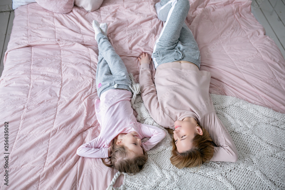 Beautiful young mother and her little daughter are lying together on the bed in the bedroom, playing, hugging and having fun. Maternal care and love. Horizontal photo Beautiful young mother and her little daughter are lying together on the bed in the bedroom, playing, hugging and having fun. Maternal care and love. Horizontal photo