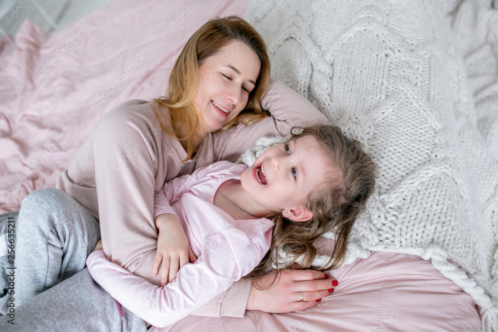 Beautiful young mother and her little daughter are lying together on the bed in the bedroom, laughing, hugging and having fun. Maternal care and love. Horizontal photo Beautiful young mother and her little daughter are lying together on the bed in the bedroom, laughing, hugging and having fun. Maternal care and love. Horizontal photo