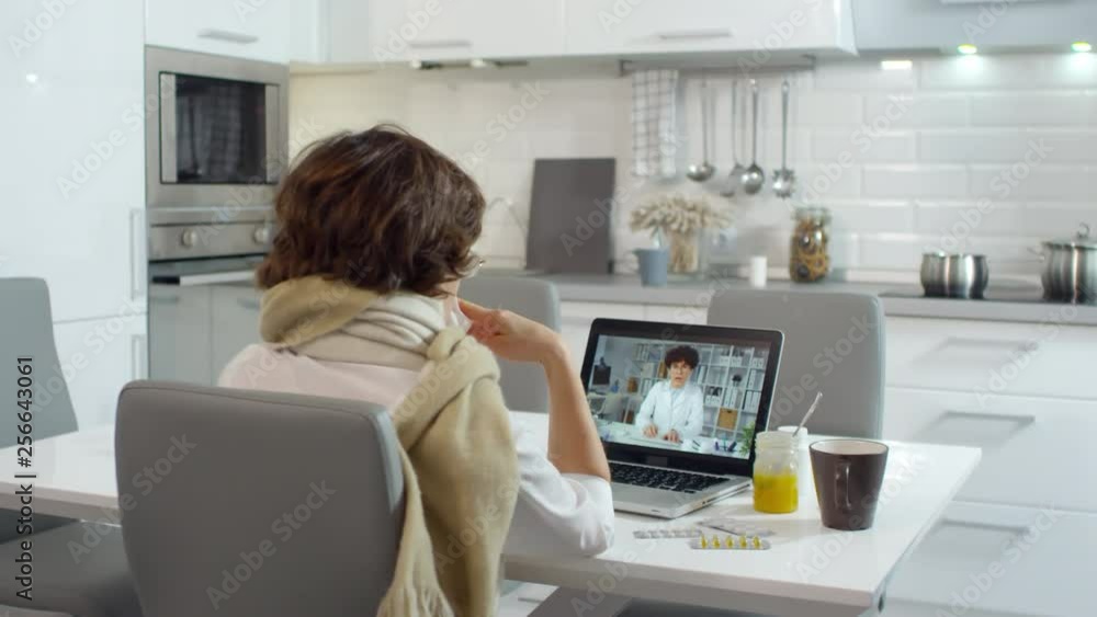 Medium shot of ill Caucasian woman sitting at PC and talking to female doctor about her health state via internet