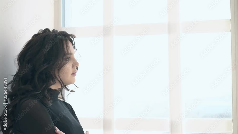 attractive young lady with large grey eyes wearing black clothes looks out window and turns to camera in room attractive young lady with large grey eyes wearing black clothes looks out window and turns to camera in room