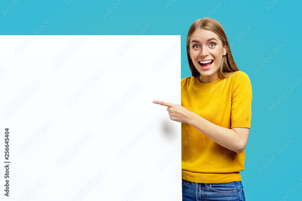 Happy girl with white empty board, pointing away, on blue background. Copy space for your text. Happy girl with white empty board, pointing away, on blue background. Copy space for your text.