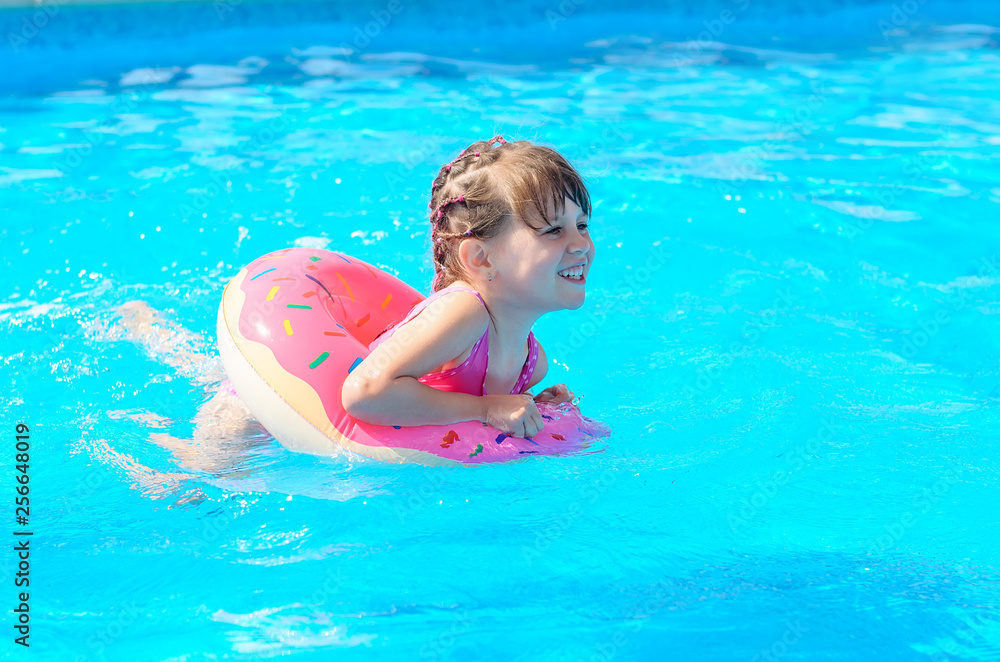 Cute smiling little girl with inflatable donut circle learns to swim in swimming pool on hot sunny day. Healthy and happy childhood concept.