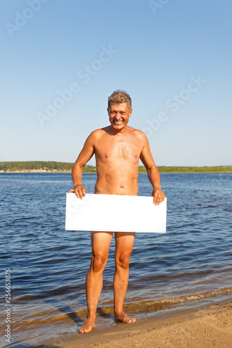 A smiling mature man stands on the beach of the river and holds a white rectangle in his hands, a poster.