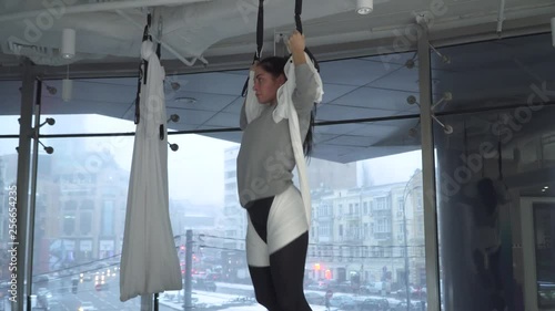 Beautiful woman hovers in hammock for yoga takes different poses in studio indoors. ityscape behind floor-to-ceiling window. Athletic girl practicing aerial yoga. Camera approaches and moves away photo