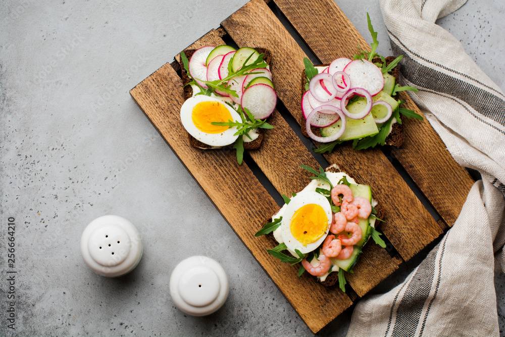 Open sandwiches on dark rye bread with eggs, shrimps, radishes, cucumber, cream cheese and arugula for breakfast on old dark background. Smorrebrod Traditional dish of Danish cuisine. Top view. Open sandwiches on dark rye bread with eggs, shrimps, radishes, cucumber, cream cheese and arugula for breakfast on old dark background. Smorrebrod Traditional dish of Danish cuisine. Top view.