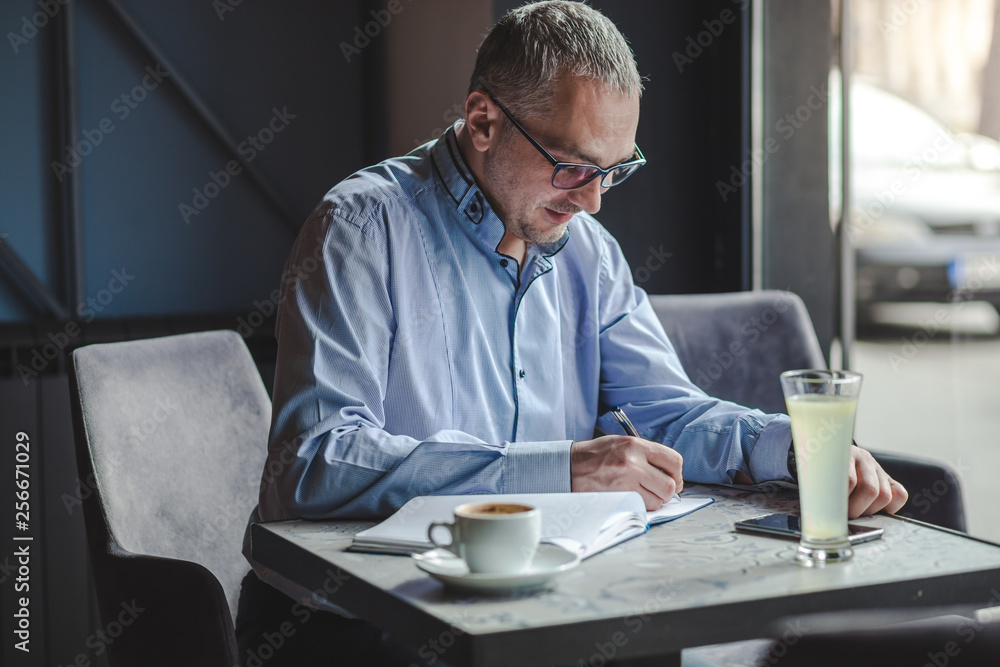 Caucasian middle-aged businessman writing down something in agenda while sitting in cafe