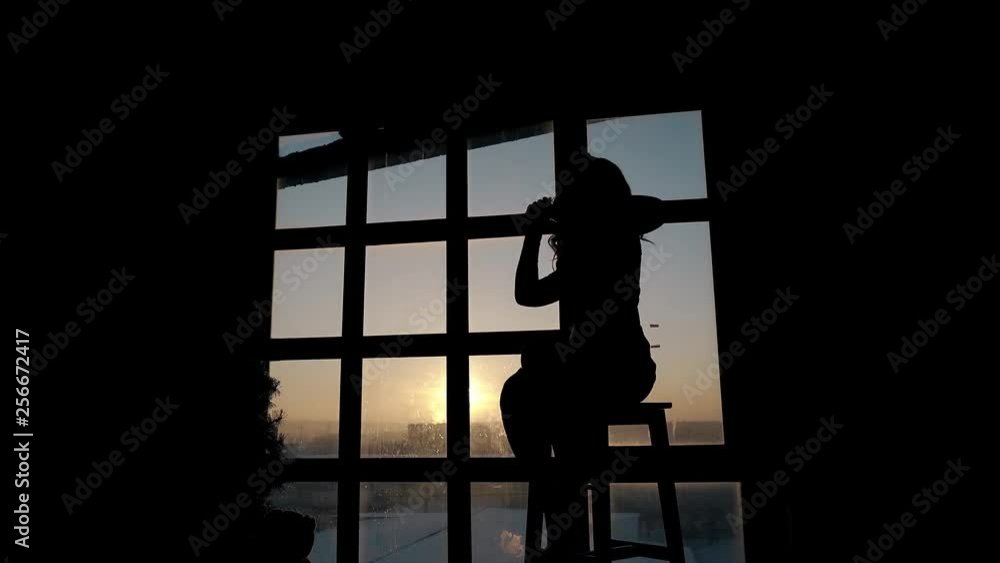 young elegant woman enjoys music and dances on stool against panoramic window in dark room in evening young elegant woman enjoys music and dances on stool against panoramic window in dark room in evening