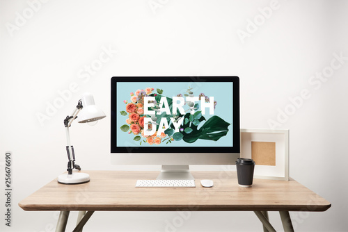 computer with green monstera leaves and flowers and earth day lettering on monitor on wooden desk