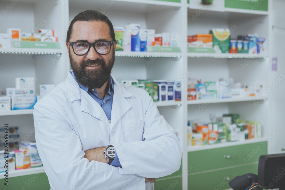 Proud mature male pharmacist smiling to the camera with arms crossed, posing at his new local drugstore. Cheerful chemist welcoming customers in his pharmacy. Sales, small business, health concept