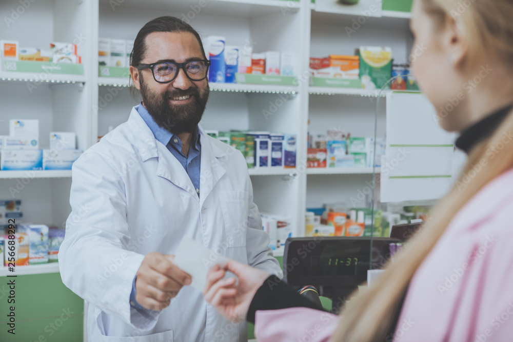 Cheerful bearded pharmacist working at the drugstore, giving prescription paper to his female customer. Unrecognizable woman buying medications at the local pharmacy. Consumerism, health concept