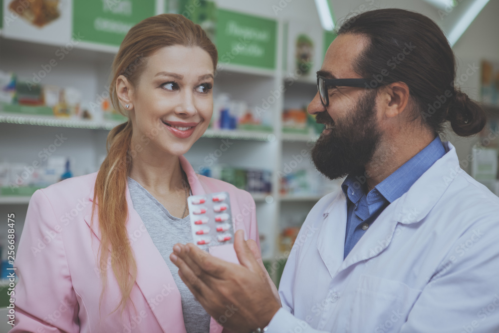 Lovely young woman smiling, buying pills at the drugstore. Bearded mature pharmacist helping his female customer showing her blister of pills. Female customer shopping for medicine at pharmacy
