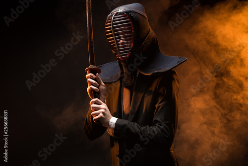 Kendo fighter in formal wear and helmet holding bamboo sword on dark