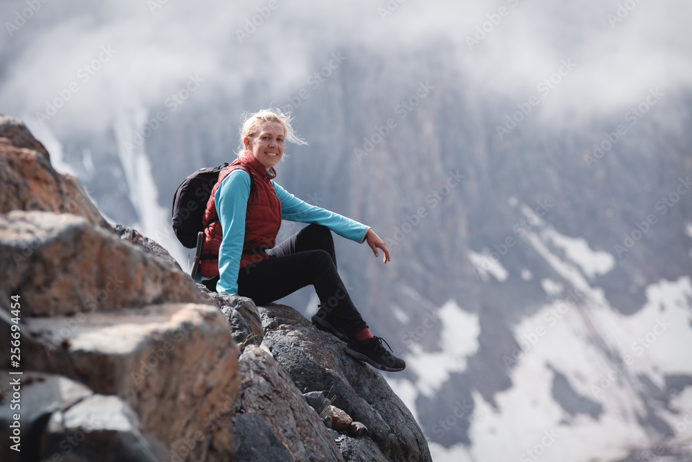 smiling blonde with a backpack sitting on a rock. the concept of traveling in mountains