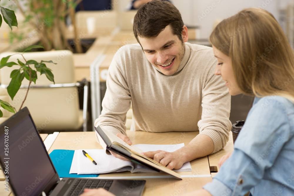 Handsome young man smiling showing something in a book to his beautiful female friend while studying together students workers creative team startup students education project colleagues interaction Handsome young man smiling showing something in a book to his beautiful female friend while studying together students workers creative team startup students education project colleagues interaction