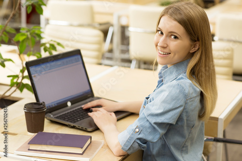 Pretty young woman browsing internet on her laptop smiling to the camera over her shoulder sitting at her workplace education working studying online internet connectivity home cafe lifestyle concept