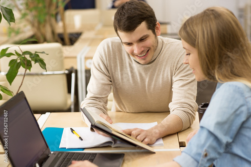 Handsome young man smiling showing something in a book to his beautiful female friend while studying together students workers creative team startup students education project colleagues interaction