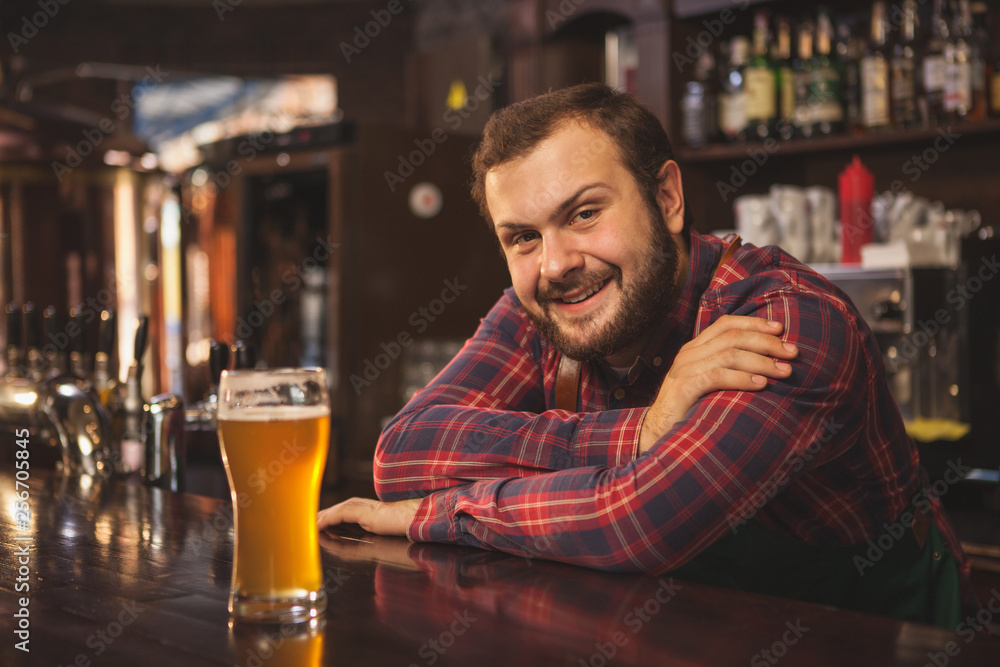 Friendly bearded bartender smiling joyfully to the camera, enjoying working at his beer pub, copy space. Happy brewer serving delicious beer, leaning on a counter. Service, lifestyle concept Friendly bearded bartender smiling joyfully to the camera, enjoying working at his beer pub, copy space. Happy brewer serving delicious beer, leaning on a counter. Service, lifestyle concept