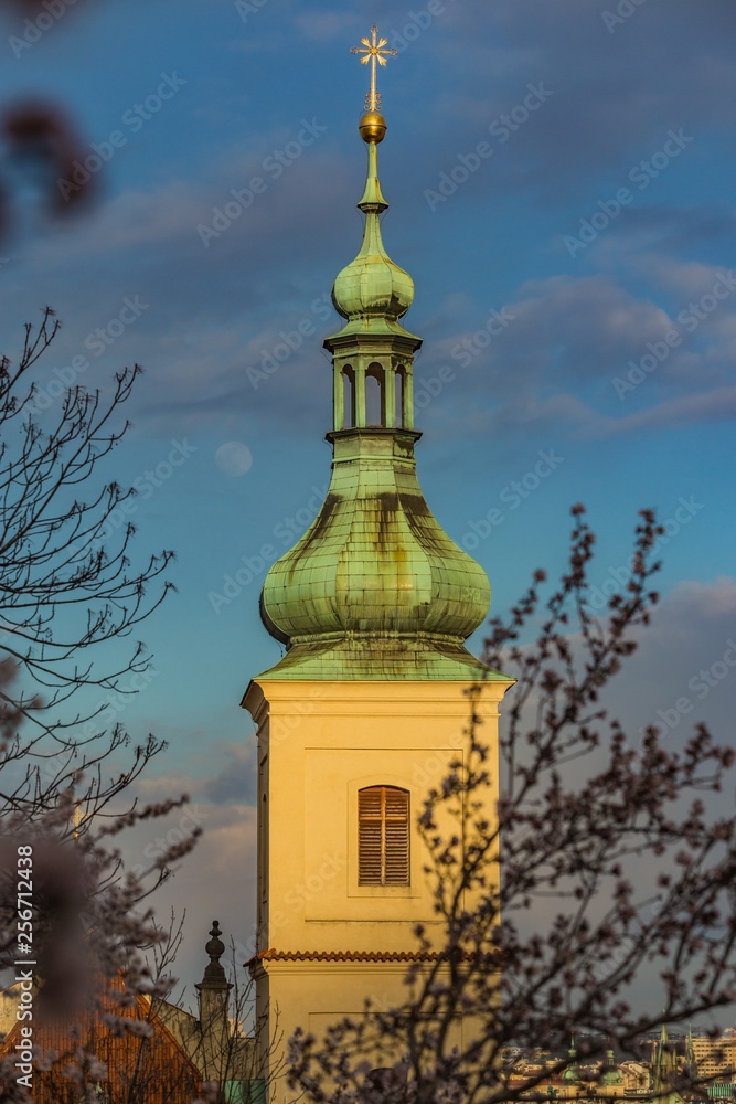Yellow bell tower with green roof and golden star, a part of the Church of Our Lady Victorious and the Prague Infant Jesus in Prague, Czech Republic, sunny spring day, blue sky, white moon, blossom