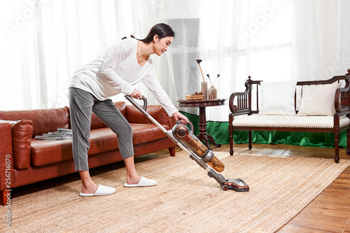 Asian teenage girl using a wireless vacuum cleaner to clean the carpet in the living room in the living room
