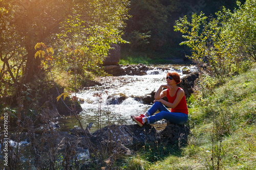 Young woman in sunglasses sits on a stone near a stream on a sunny autumn day