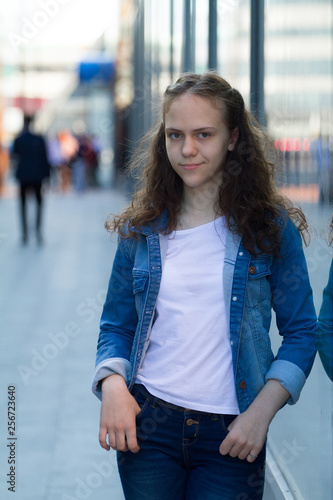 beautiful teen girl in denim clothes stands leaning on a glass building on the city street.