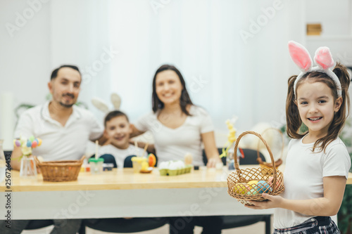 Pretty small girl with pigtails and bunny ears standing on the forefront with the basket of colored eggs in her hands. Her father mother and brother sitting at the table, preparing for Easter.