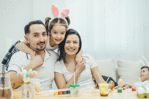 Happy lovely girl with bunny ears hugging her smiling parents on the forefront. Little son is sleeping on the sofa on the blurred background.