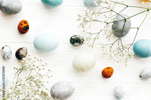 Stylish Easter eggs with spring flowers flat lay on white wooden background. Modern easter eggs painted with natural dye in yellow,blue,green,grey colors. Happy Easter greeting card