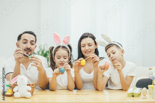 Inspired family sitting together at the table  brother and sister in bunny ears. Everyone is painting different easter eggs with different colors.