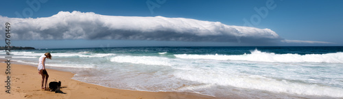 Girl on beach with dog unaware of approaching thunderstorm, a rare dramatic coastal Volutus roll cloud, a form of shelf cloud, out to sea. Culburra Bach, NSW Australia