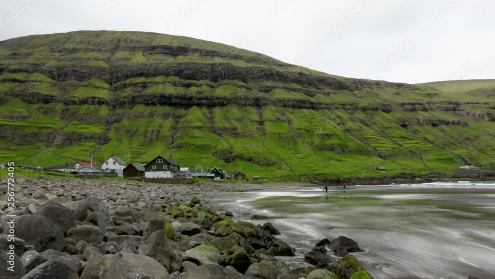 People walking on black sandy beach in Tjornuvik with village and huge green mountain wall in background People walking on black sandy beach in Tjornuvik with village and huge green mountain wall in background