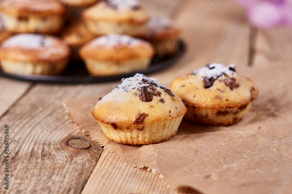 The two cupcakes with chocolate chips with powdered sugar lie on a kraft paper next to other cupcakes in a black baking sheet and orchid flower on a wooden background. Close-up The two cupcakes with chocolate chips with powdered sugar lie on a kraft paper next to other cupcakes in a black baking sheet and orchid flower on a wooden background. Close-up