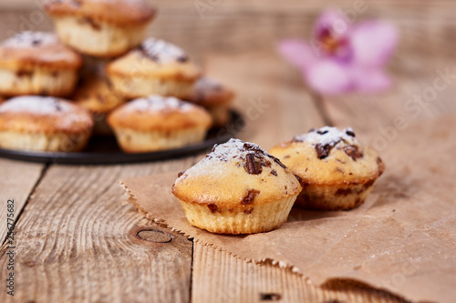 The two cupcakes with chocolate chips with powdered sugar lie on a kraft paper next to other cupcakes in a black baking sheet and orchid flower on a wooden background.