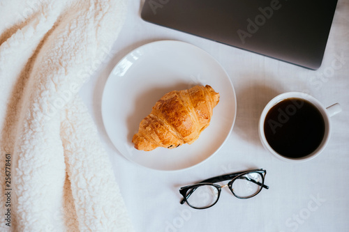 Big white morning cup of coffee and crossant in bed with notebook, glasses, pillow and cosy blanket. photo