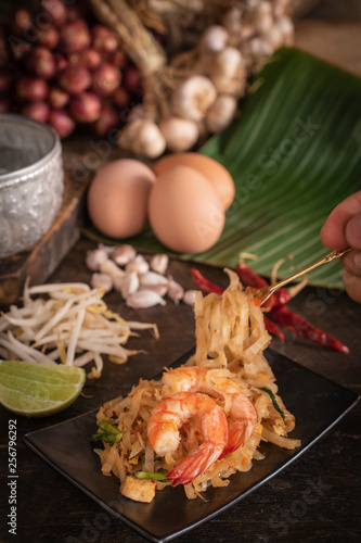 Thai fried noodles or pad thai with shrimp on black plate placed on the wood table there are eggs, garlic, bean sprouts, shallot, silver water bowl, chilli and cutting board placed back side. 
